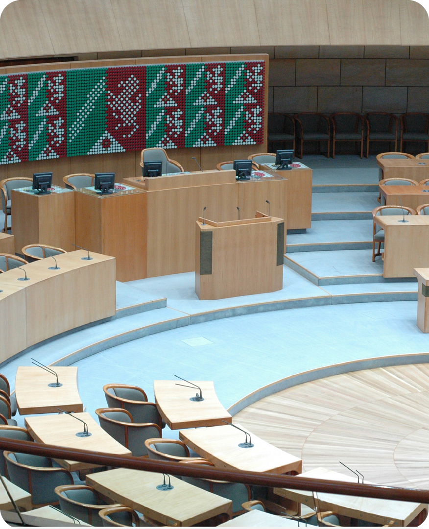 Innenansicht des Plenarsaals des Landtags Nordrhein-Westfalen in Düsseldorf: Halbkreisförmig angeordnete Sitzreihen, Rednerpult im Zentrum und die Landesfarben rot, weiß und grün als Wandgestaltung.